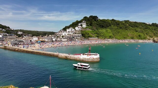 boat passing by the pier of Looe beach, people on the beach, and coastal buildings in Cornwall, UK