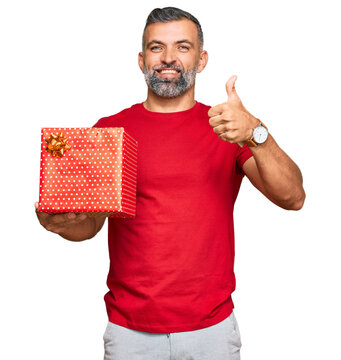 Middle age handsome man holding gift smiling happy and positive, thumb up doing excellent and approval sign