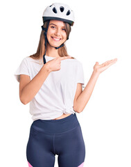 Young beautiful girl wearing bike helmet amazed and smiling to the camera while presenting with hand and pointing with finger.