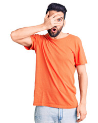 Young handsome man with beard wearing casual t-shirt peeking in shock covering face and eyes with hand, looking through fingers with embarrassed expression.