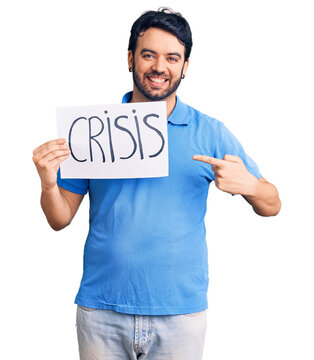 Young hispanic man holding crisis message paper smiling happy pointing with hand and finger
