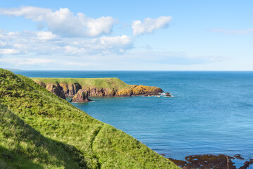 View of a rugged coast in Scotland on a sunny summer day. A fishing boat is visible in distance.