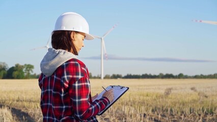 Woman engineer wearing a white protective helmet is taking notes with a clipboard in a field with wind turbines, as the sun sets. Clean energy and engineering concept