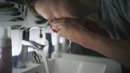 Caucasian boy stands at sink in bathroom, washes face with cold water and wipes hands using towel. Teenager looks at his reflection in the mirror and makes morning bathroom routine. Low angle view.