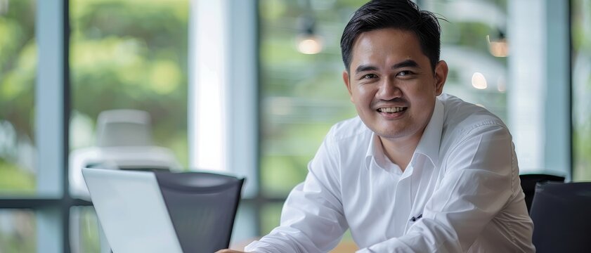 A relaxed professional in white shirt sits in an airy office, smiling confidently at the camera, a laptop visible on the desk.