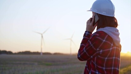 Woman engineer talking on the phone in a field with wind turbines, as the sun sets. Concept of clean energy and engineering