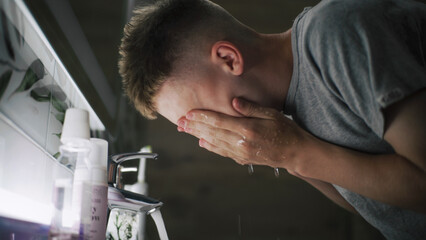 Caucasian boy comes to sink in bathroom, washes face with cold water and looks at his reflection in...