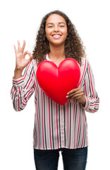 Young hispanic woman in love holding red heart doing ok sign with fingers, excellent symbol