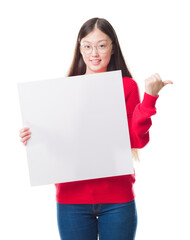 Young Chinese woman over isolated background holding blank banner pointing and showing with thumb...