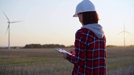 Adult woman engineer wearing white cask is taking notes on a clipboard on a field with wind turbines