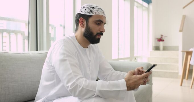 Focused young Arab man in white kandura dress and skullcap typing on smartphone, chatting on Internet, texting message, using gadget for online communication, getting happy