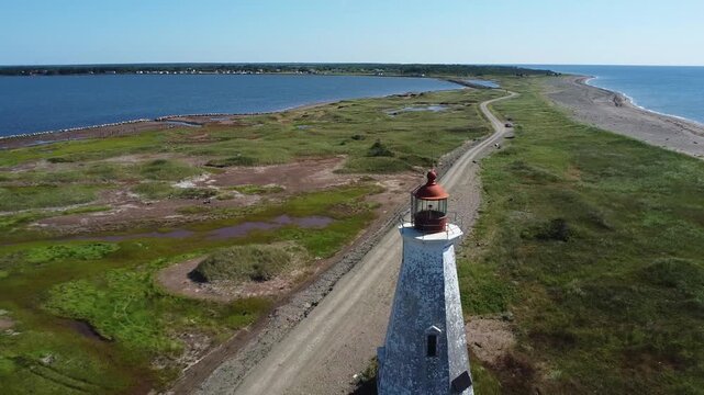 Un drone s'&eacute;loigne du vieux phare Chiasson-Office, datant de 1872, Ile Lam&egrave;que. Une ancien phare rouge & blanc sur une pointe de de sable mar&eacute;cageuse sous un ciel bleu sans nuage au Nouveau-Brunswick.