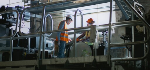 Two professional heavy industry engineers in protective uniforms and helmets at manufacturing facility or factory. Male worker discusses production and equipment with mature supervisor. Slow motion.