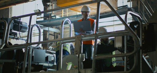 Professional heavy industry mature engineer in safety uniform, helmet and goggles uses laptop and talks with technician at manufacturing factory. Two male employees work with equipment. Slow motion.