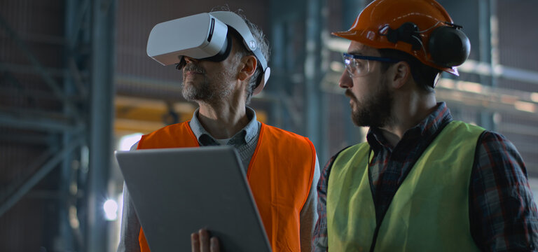Two professional industry technicians wearing safety uniform and hardhats stand at manufacturing factory, use laptop and AR headset. Mature engineer and adult employee work in warehouse. Slow motion.