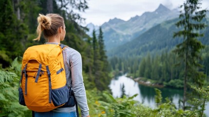 Naklejka premium A woman with a blond bun wearing a bright orange backpack stands near a river, looking towards rugged mountains and dense forest, showcasing outdoor adventure and tranquility.