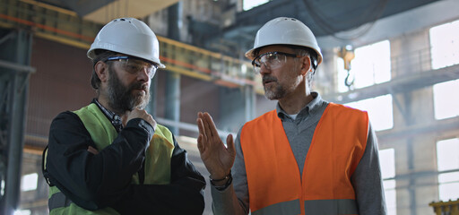 Two professional heavy industry engineers in safety uniform and hard hats discuss work process at manufacturing factory. Mature supervisor talks to adult worker at plant or workshop. Slow motion.