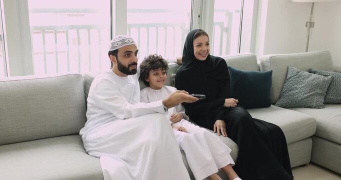 Positive young Arab parents and little son kid watching movie, sitcom show, cartoon on TV, using remote control, pushing button, sitting on home sofa, laughing, enjoying leisure