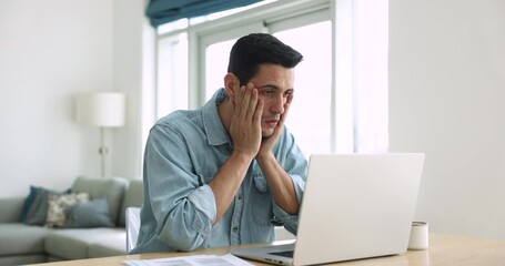 Serious young Latin freelancer employee man working at laptop from home, getting shocking bad news, getting sad, frustrated, confused, touching head in despair, staring at computer screen - Powered by Adobe