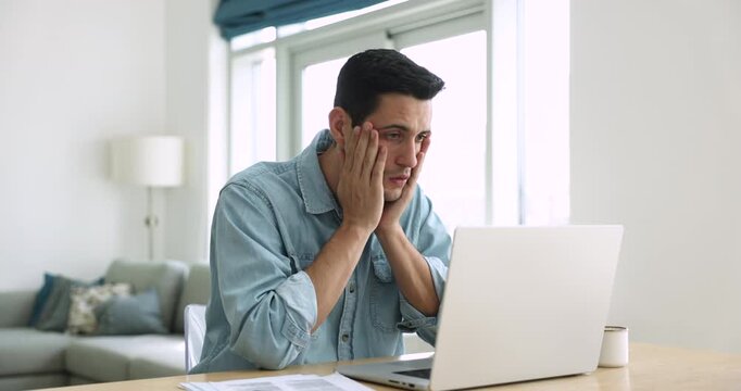 Serious young Latin freelancer employee man working at laptop from home, getting shocking bad news, getting sad, frustrated, confused, touching head in despair, staring at computer screen