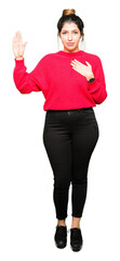 Young beautiful woman wearing red sweater and bun Swearing with hand on chest and open palm, making...