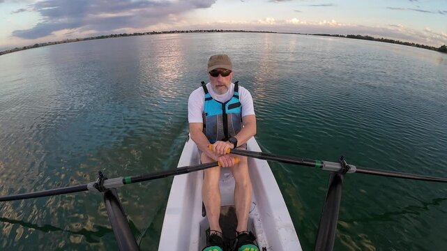 senior man in a rowing shell on a calm lake at evening, POV from a boat stern