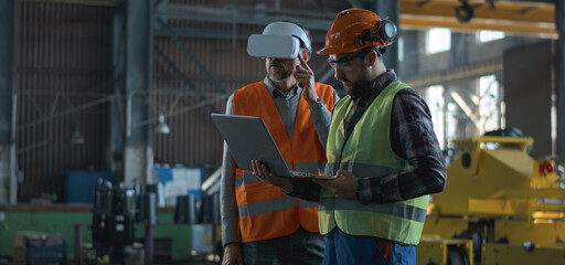 Two professional industry technicians wearing protective uniform and helmets stand at manufacturing factory. Mature engineer uses VR headset, adult employee work in laptop computer. Slow motion.