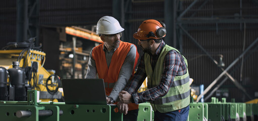 Two heavy industry engineers in uniform and helmets stand at manufacturing factory, work using...