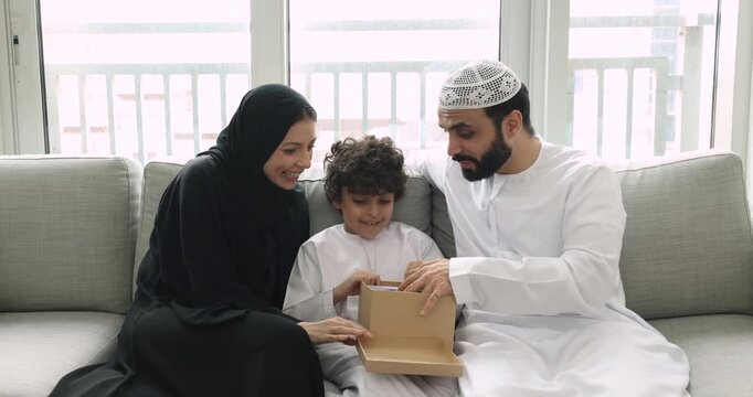 Happy Arabic couple of parents and adorable son boy in traditional Muslim clothes opening package at home, getting purchase from webstore, unpacking parcel delivered by post service