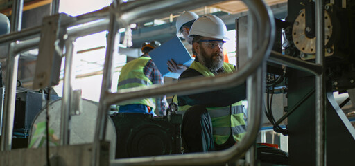 Two professional heavy industry technicians in safety uniforms and hard hats work with equipment at manufacturing factory. Mature supervisor or engineer uses laptop and talks to workers. Slow motion.