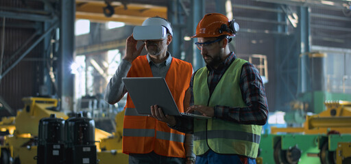 Two professional industry technicians wearing protective uniform and helmets stand at manufacturing factory. Mature engineer uses VR headset, adult employee work in laptop computer. Slow motion.