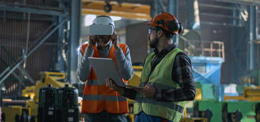 Two professional industry technicians wearing protective uniform and helmets stand at manufacturing factory. Mature engineer uses VR headset, adult employee work in laptop computer. Slow motion.