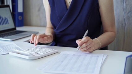 Fototapeta premium Businesswoman with dark blue dress is using white calculator and taking notes in her office. Taxes and audit in business