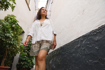 Confident woman walking along a narrow alleyway, wearing a casual white shirt and patterned shorts, exuding a sense of freedom and joy