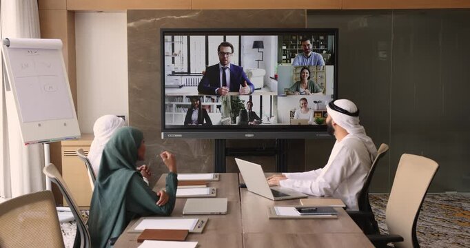 Business leader man holding online meeting with diverse Arabic and European colleagues, speaking from large split screen, talking to Muslim team at office table and freelance partners on display