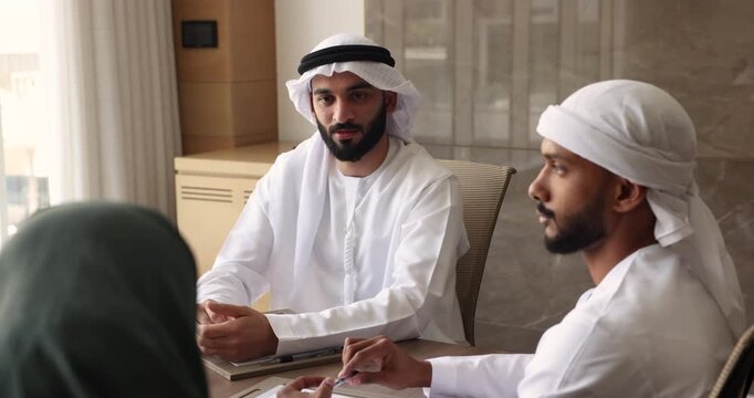 Young male Arab boss talking to employees in Muslim clothes, sitting at meeting table, speaking to colleagues, partners. Arabic businesspeople discussing teamwork, cooperation