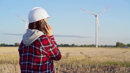 Woman engineer is talking on a smartphone in a field with wind turbines as the sun sets. Concept of clean energy and engineering audit