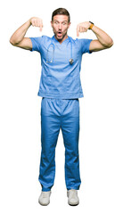 Handsome doctor man wearing medical uniform over isolated background Pointing down with fingers showing advertisement, surprised face and open mouth