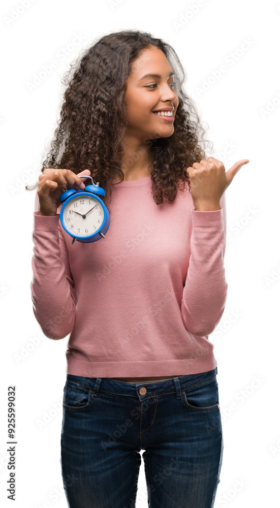 Wall mural Young hispanic woman holding alarm clock pointing and showing with thumb up to the side with happy face smiling