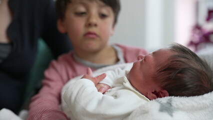 Young boy gently holding newborn sibling, with mother close by, exploring family dynamics, nurturing, and sibling bond, cozy indoor scene, emotional connection