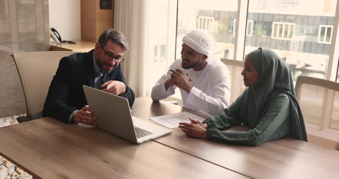 Positive European business professional man in suit showing online presentation on laptop to young Arabic couple, selling real estate property for investment, meeting with clients in office