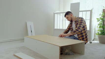 Man sitting down on the carpet takes apart a white wooden furniture and removes the screws from the furniture with a tool. Daddy disassembles a bouncer from the child's old crib. Furniture assembly - Powered by Adobe