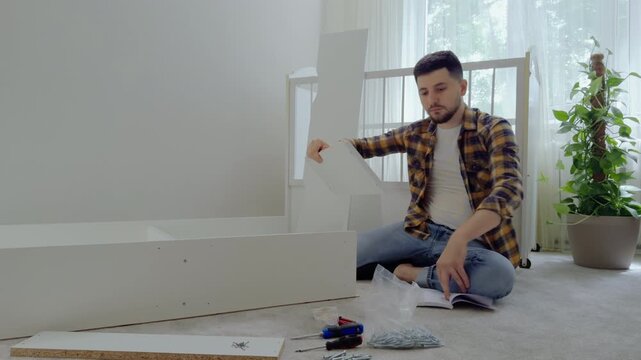 Young man prepares the future child's room by assembling the furniture. A bright and minimalist room for the baby. Dad assembles the crib for the newborn with the help of the instruction book