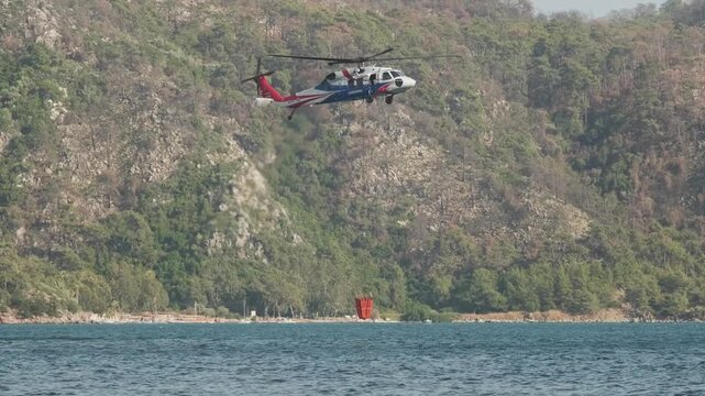 A helicopter hovers low over a lake, drawing water into its bucket to aid in firefighting efforts, with dense forested mountains in the background.
