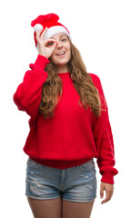 Young blonde woman wearing santa claus hat with happy face smiling doing ok sign with hand on eye looking through fingers