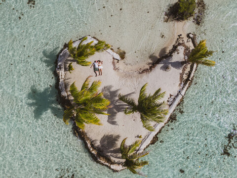 Drone view looking down on a man and woman laying on white sand on a heart shaped island in the south pacific. 