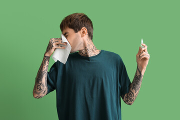 Allergic young man with nasal drops and tissues on green background