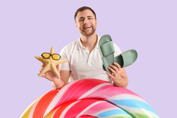 Portrait of young man with inflatable mattress, flip-flops and starfish on lilac background