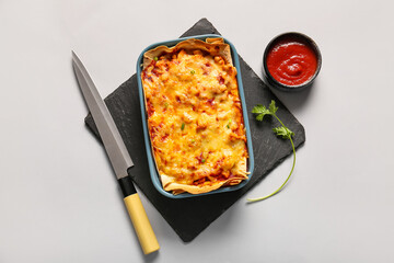 Baking dish with tasty enchilada, knife and tomato sauce on light background