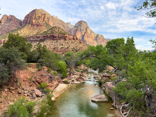 Slow moving river among red rocks and greenery, with large Zion Park mountains frame left in the background